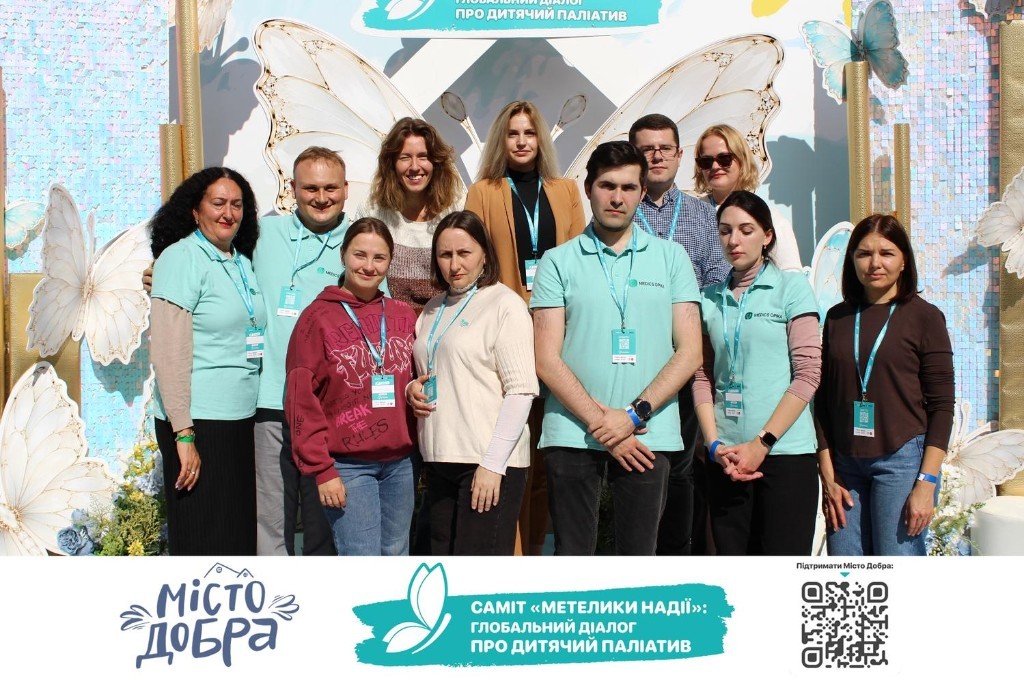 Команда учасників саміту «Метелики Надії» на тлі банера про дитячий паліатив у «Місті Добра»